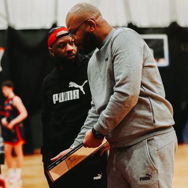 Two basketball coaches discuss strategy during a game, one holding a clipboard.