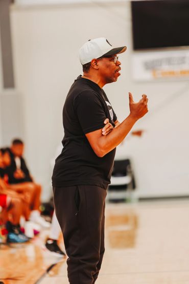 Man in black sportswear and cap speaking passionately on indoor basketball court.