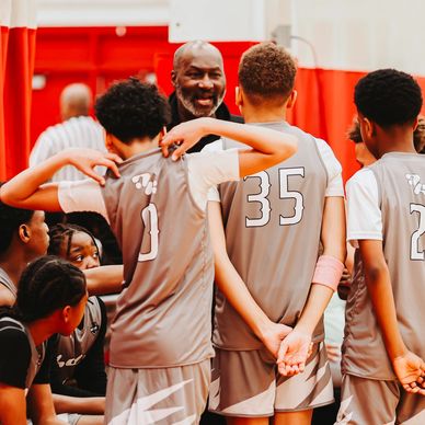 Youth basketball team gathers around coach for a timeout in a gym.