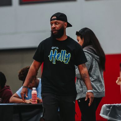 Man in black cap and t-shirt walking on a basketball court with people in the background.