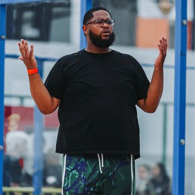 Man in sports attire gesturing on indoor basketball court, kids nearby.
