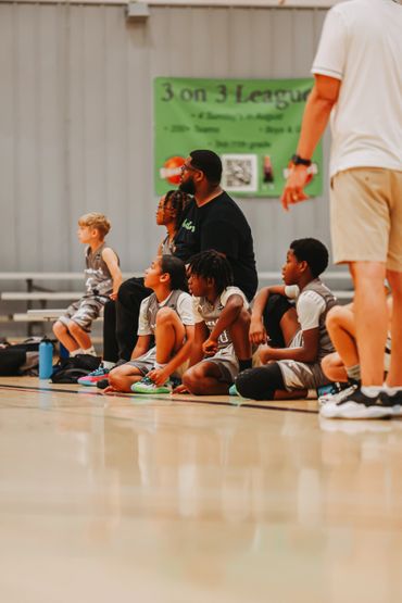 Young basketball players and coach watching a game from the sidelines in a gym.