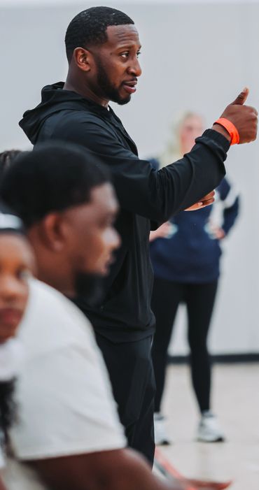 Man in black hoodie gestures with thumb up during a conversation indoors.