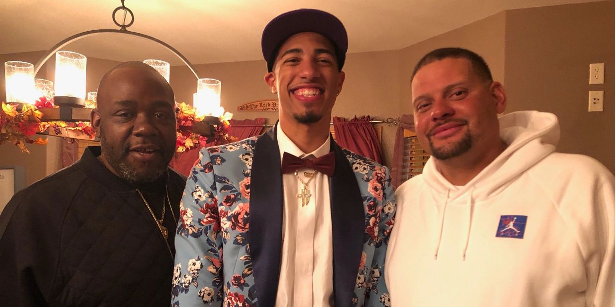 Three men smiling indoors, one in a floral suit and bow tie.