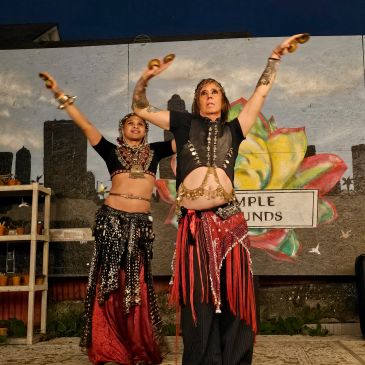 Two women in traditional belly dance costumes perform with raised arms outdoors.