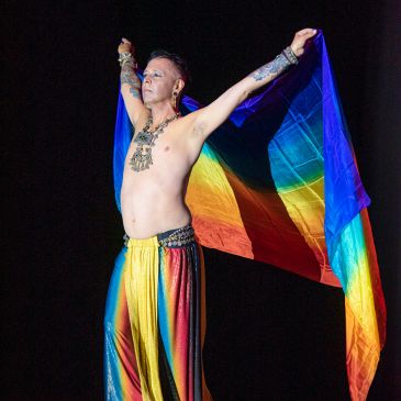 Performer holding a rainbow flag wearing colorful pants and jewelry.