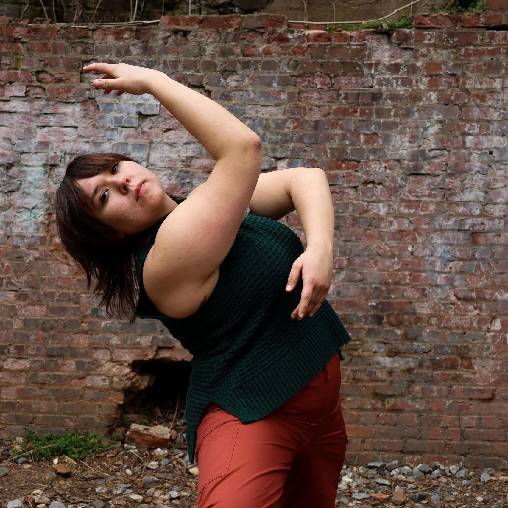 Cecilia wears a dark green knit tank top and rust colored pants. She is standing in a wide stance wi