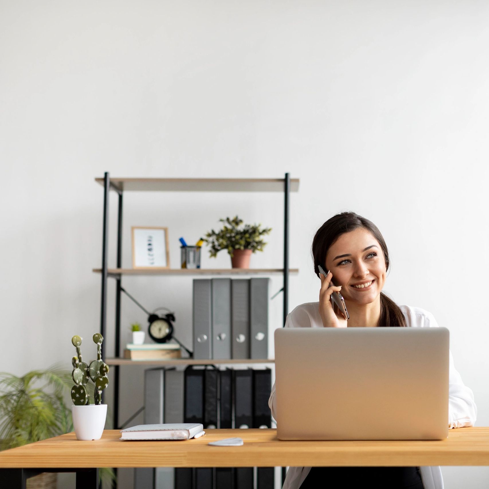 Smiling woman talking on phone at her desk with a laptop and plants.