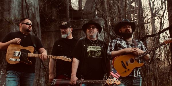 Four men with guitars in a wooded outdoor setting.