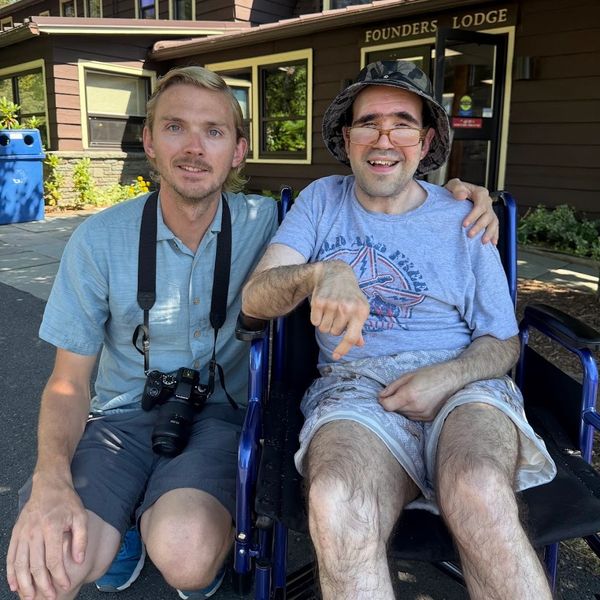 Two friends posing outdoors, one in a wheelchair.