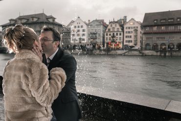 Couple sharing a kiss in the rain by a river with old buildings in the background.