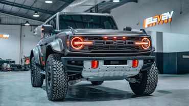 A customized matte black Ford Bronco with glowing orange headlights in a garage.