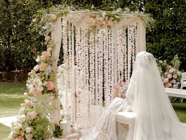 Bride in a flowing veil sitting beside a floral arch in a serene garden.