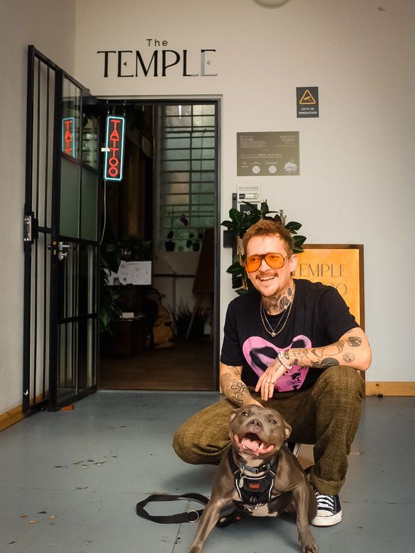 A tattooed man wearing orange sunglasses smiles while crouching next to a happy dog in front of a tattoo studio.
