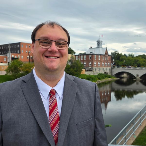 Man in suit standing by a river with buildings in the background.
