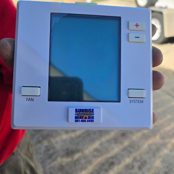 Hand holding a white HVAC thermostat with a blue screen and control buttons.