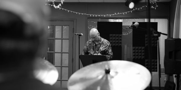 Musician playing guitar in a cozy, dimly lit studio with drum set in foreground.