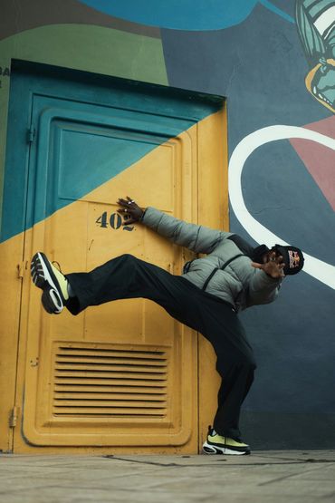 Man in a gray jacket and cap kicking in front of a colorful wall door.