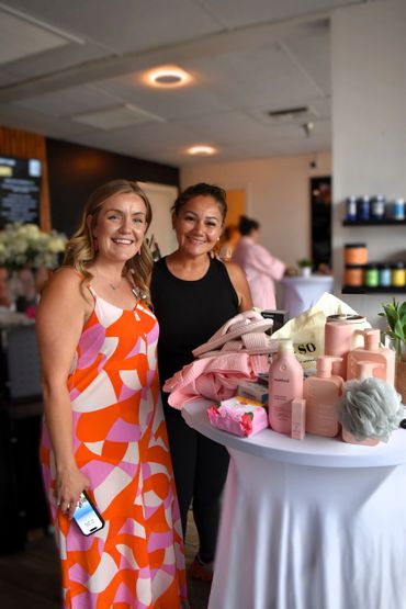 Two women smiling beside a table with pink beauty products and towels.