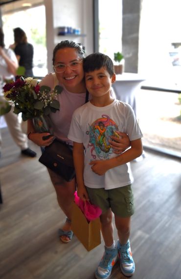 Smiling woman with flowers and a boy holding a gift bag, posing inside a bright room.
