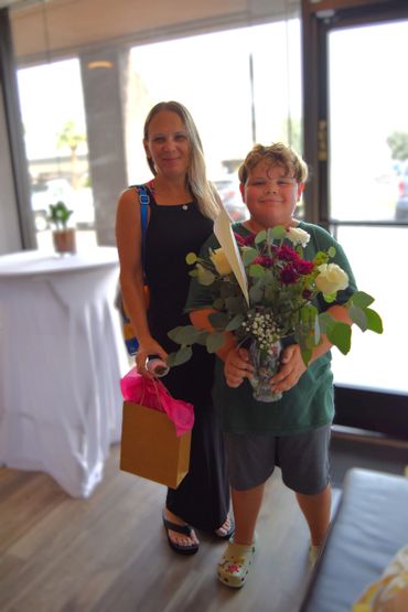A woman and a boy smiling, holding flowers and a gift bag indoors.