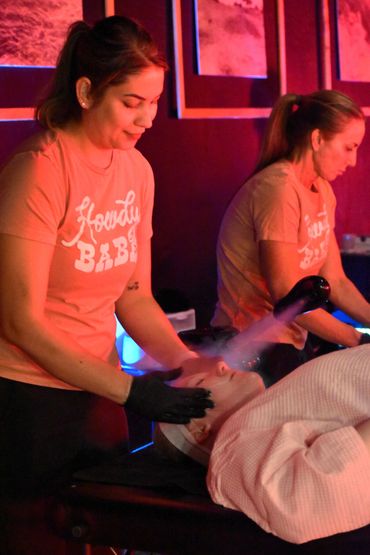 Two women perform facial treatments with steamers in a dimly lit spa.