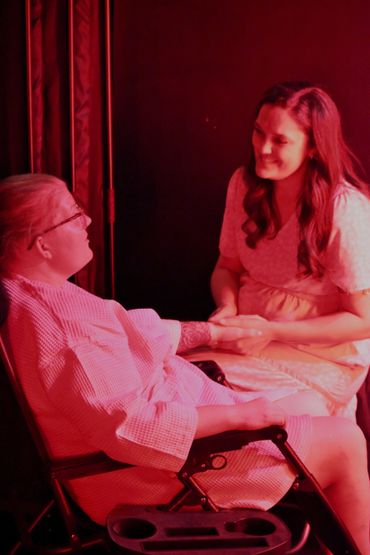 Two women share a tender moment in warm red lighting, one seated and the other holding her hand.