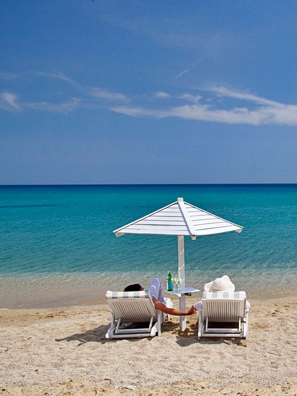 Two people relaxing on beach chairs under a white umbrella by the clear blue sea.