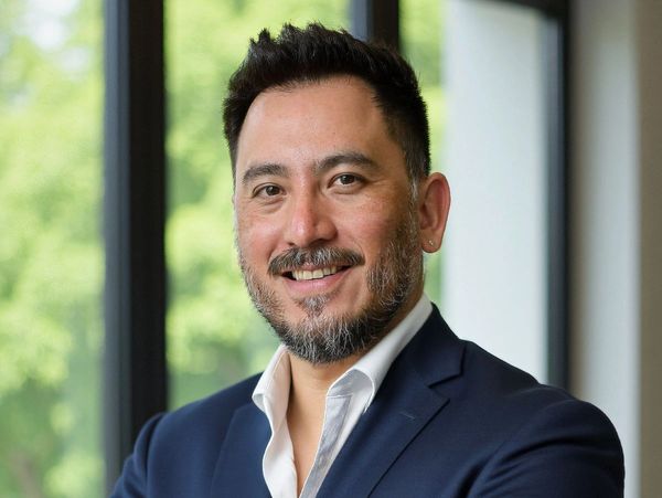 Smiling man in a navy suit with a beard standing indoors near windows.
