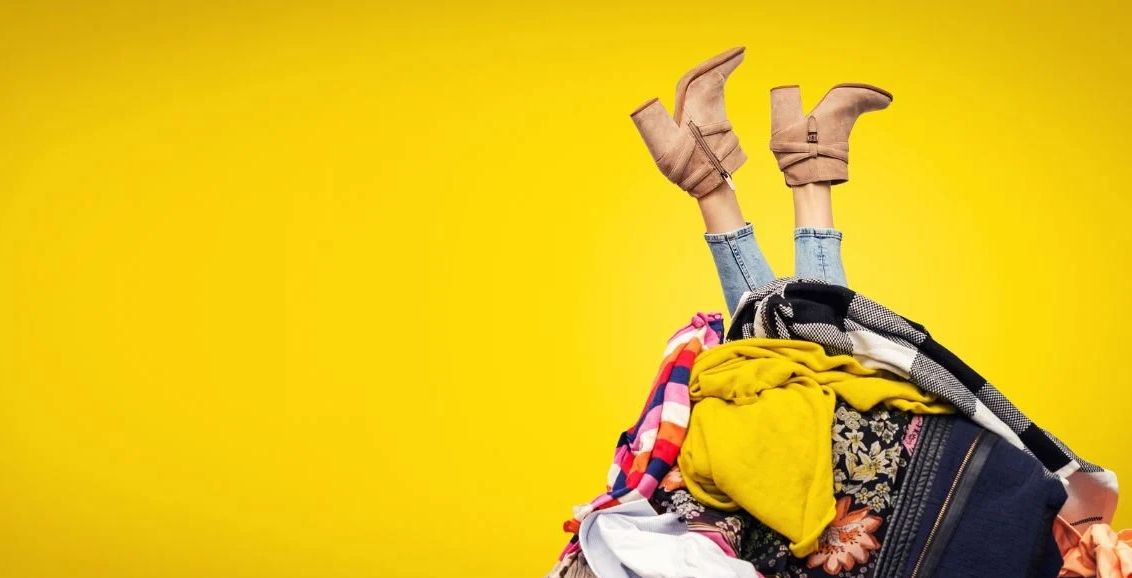 Person's legs sticking out from a large pile of colorful clothes against a yellow background.