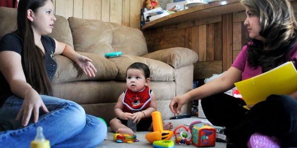 A baby sits on the floor with two adults talking nearby.