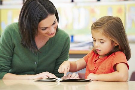 A woman helps a young girl read a book.