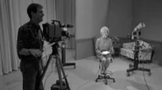 A cameraman films an older woman seated on a stool in a black-and-white television studio set.