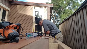 A man in a cap working on a wooden deck with tools around him.