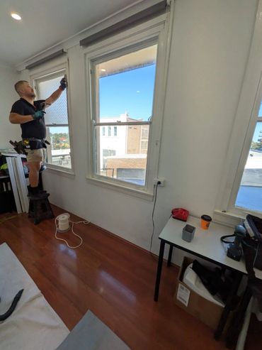 Worker installing a window screen while standing on a stool inside a room.