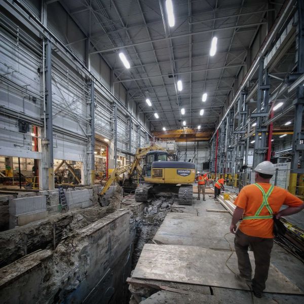 An excavator digging a hole in a manufacturing facility with construction workers looking on.