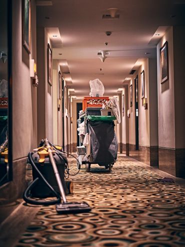 A cleaning cart and vacuum cleaner in a hotel hallway with patterned carpet.