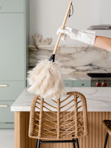 A hand with white gloves dusting a wicker chair with a feather duster in a modern kitchen.