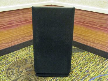 A black speaker placed in the corner of a room with patterned carpet and textured walls.