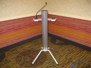 Metal podium stand with microphone in a corner room with patterned walls and carpet.