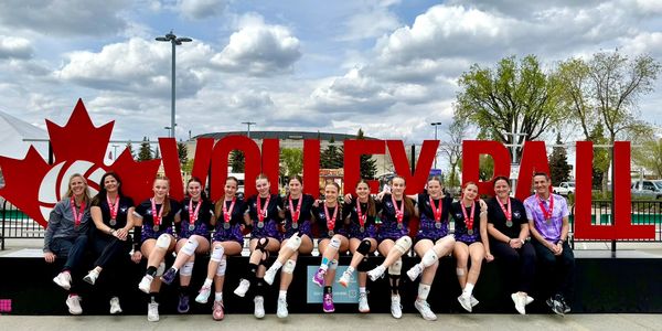 Youth volleyball team wearing medals sitting in front of large red volleyball sign outdoors.