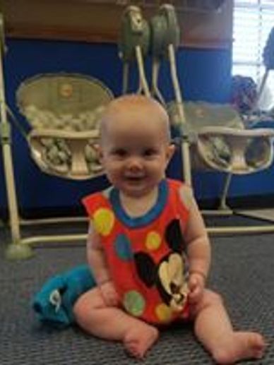 Smiling baby in colorful outfit sitting on the floor.