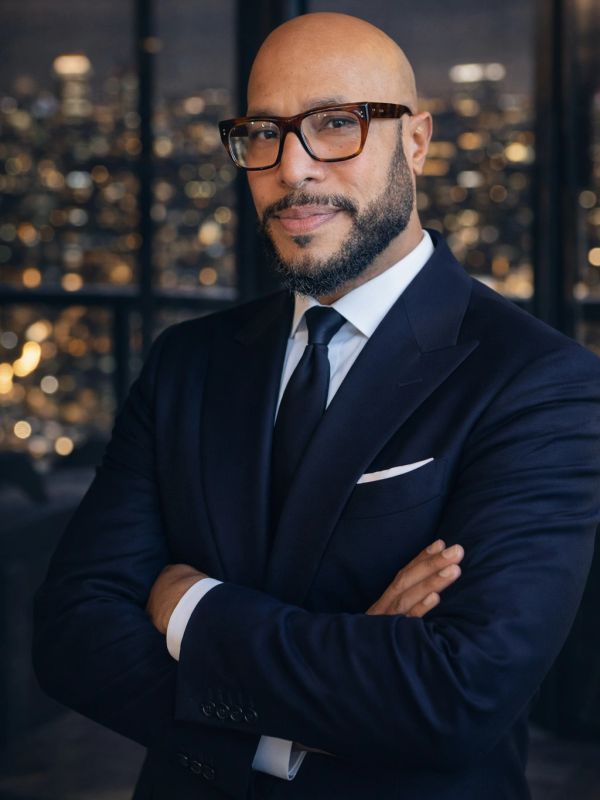 Confident bald man in glasses and suit with city lights background.