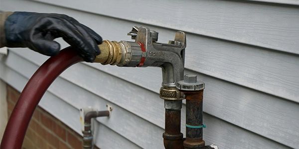 Hand in black glove attaching a hose to an outdoor faucet on a house.