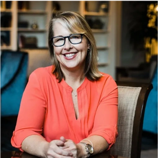 Headshot of a blonde woman with black glasses smiling and her hands resting on a table.