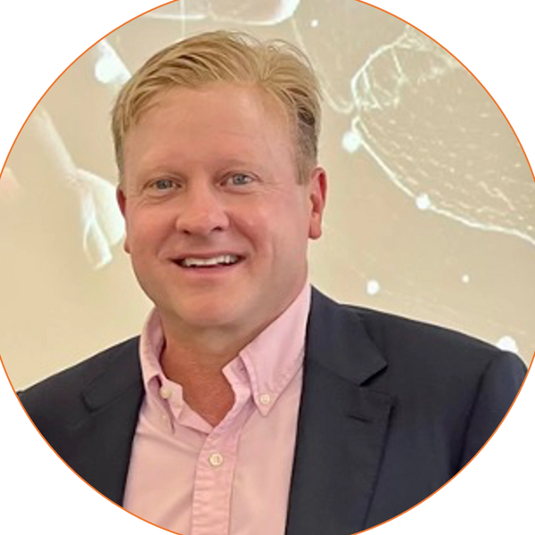 Headshot of a blonde man in his 50s smiling. He is wearing a pink dress shirt and black blazer.