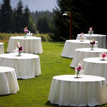 Outdoor event tables with white tablecloths and floral centerpieces on grass.