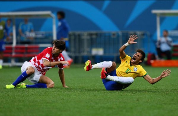 Two soccer players fall on the field during a match, one in yellow and one in red.