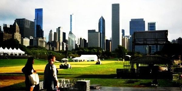Two women, stage with production crews setting up festival, green lawn with skyscrapers