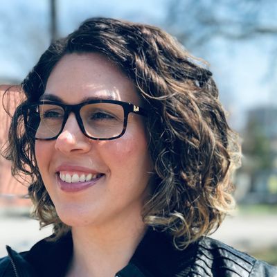 Smiling woman with curly hair and glasses outdoors on a sunny day.